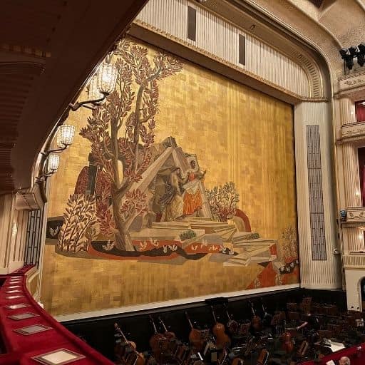 Golden stage curtain at the Vienna State Opera featuring a stylized modernist landscape scene, viewed from the red-velvet balcony with the orchestra pit below.