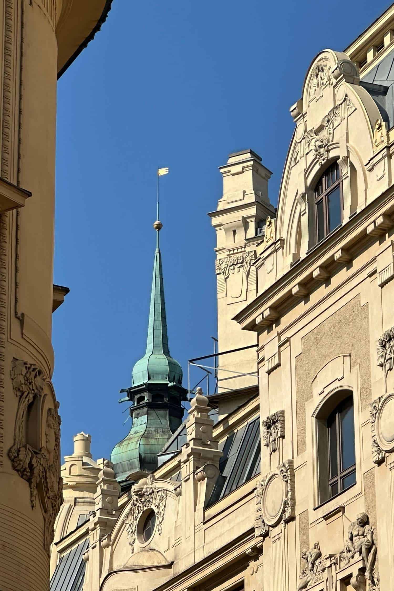 Art Nouveau architecture and historic rooftops in Prague Ornate Art Nouveau building facades in central Prague, with sculptural details and a green copper spire rising above the rooftops against a clear blue sky