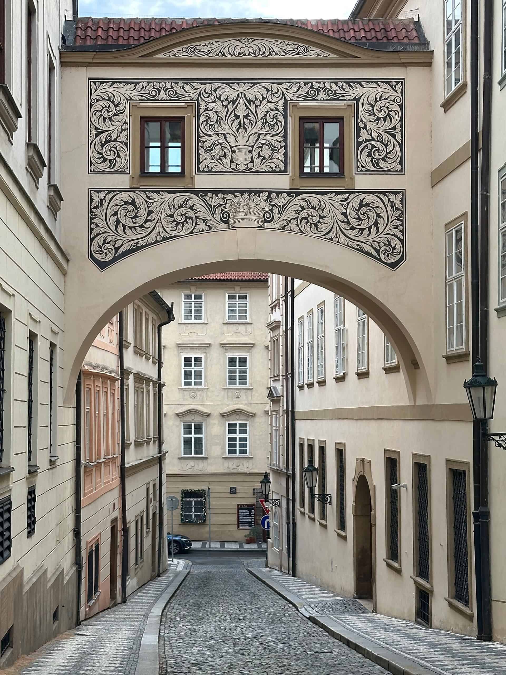 Painted Baroque archway spanning a cobblestone street in Malá Strana, Prague, with sgraffito decoration and historic townhouses beyond.