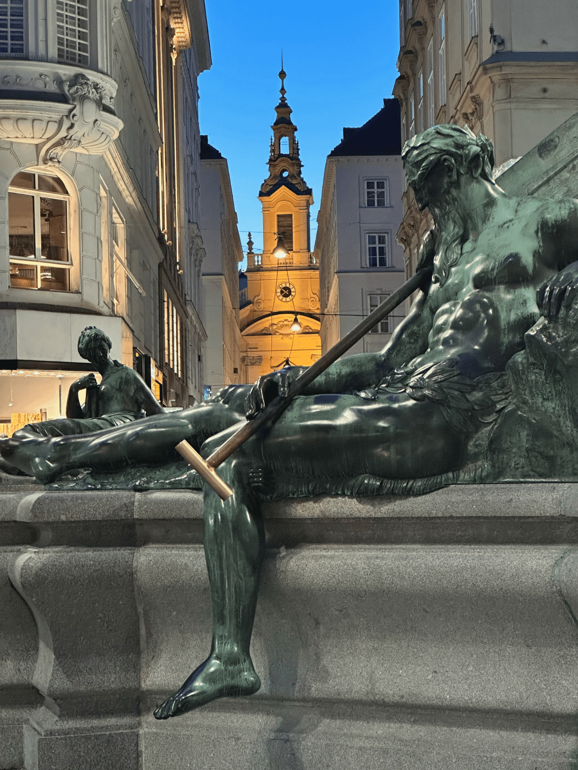 Detail of the Donnerbrunnen fountain on Neuer Markt, Vienna Bronze reclining male figures from the Donnerbrunnen fountain on Vienna’s Neuer Markt, illuminated at dusk with historic buildings in the background.