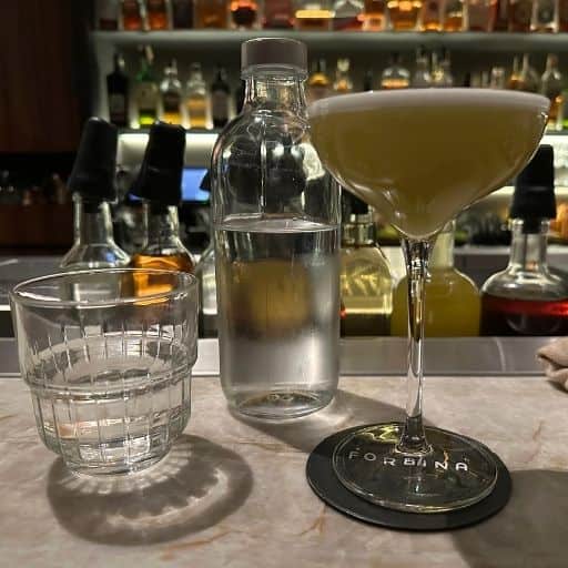 A minimalist cocktail presentation at the Forbína bar at Café Slavia in Prague, featuring a stemmed glass, water carafe, and softly lit spirits behind the bar.