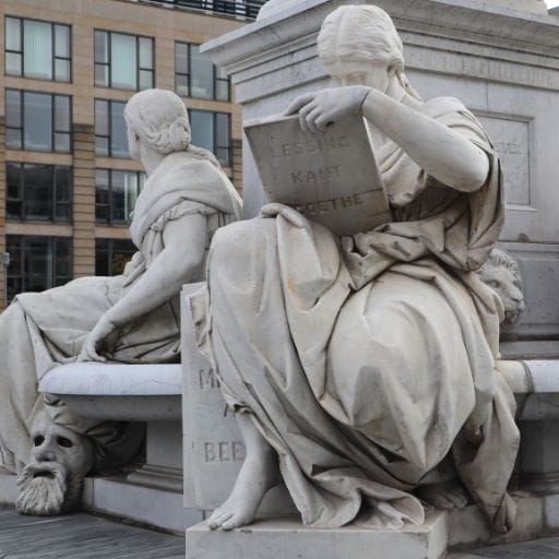 Marble allegorical figures on the Friedrich Schiller Monument in Berlin, including a seated woman reading a tablet inscribed with the names Lessing, Kant, and Goethe.