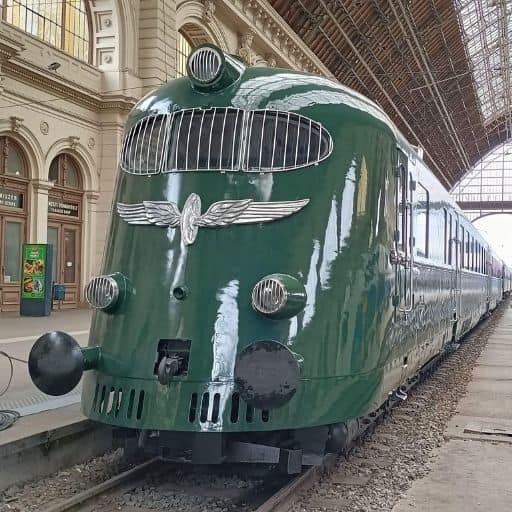 Historic streamlined green locomotive displayed inside a grand iron-and-glass railway station, reflecting early modern railway engineering and industrial design.