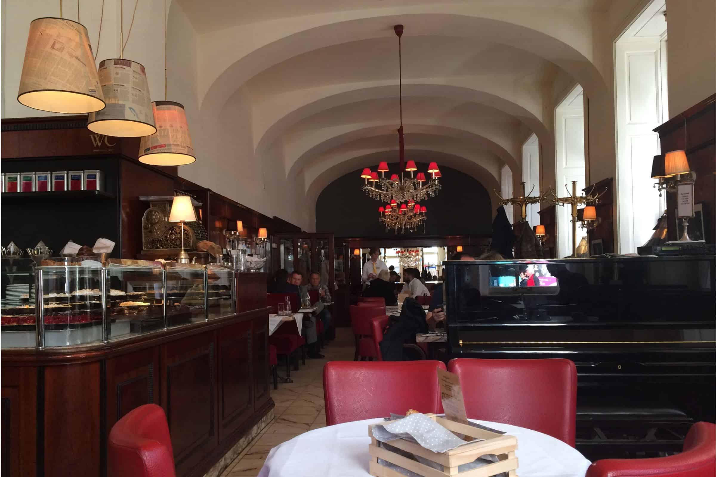 Café Diglas, Vienna — traditional coffeehouse interior with pastry counter and red leather seating Dark wood paneled interior of Vienna's Cafe Diglas with a white arched ceiling, chandeliers, and red seating