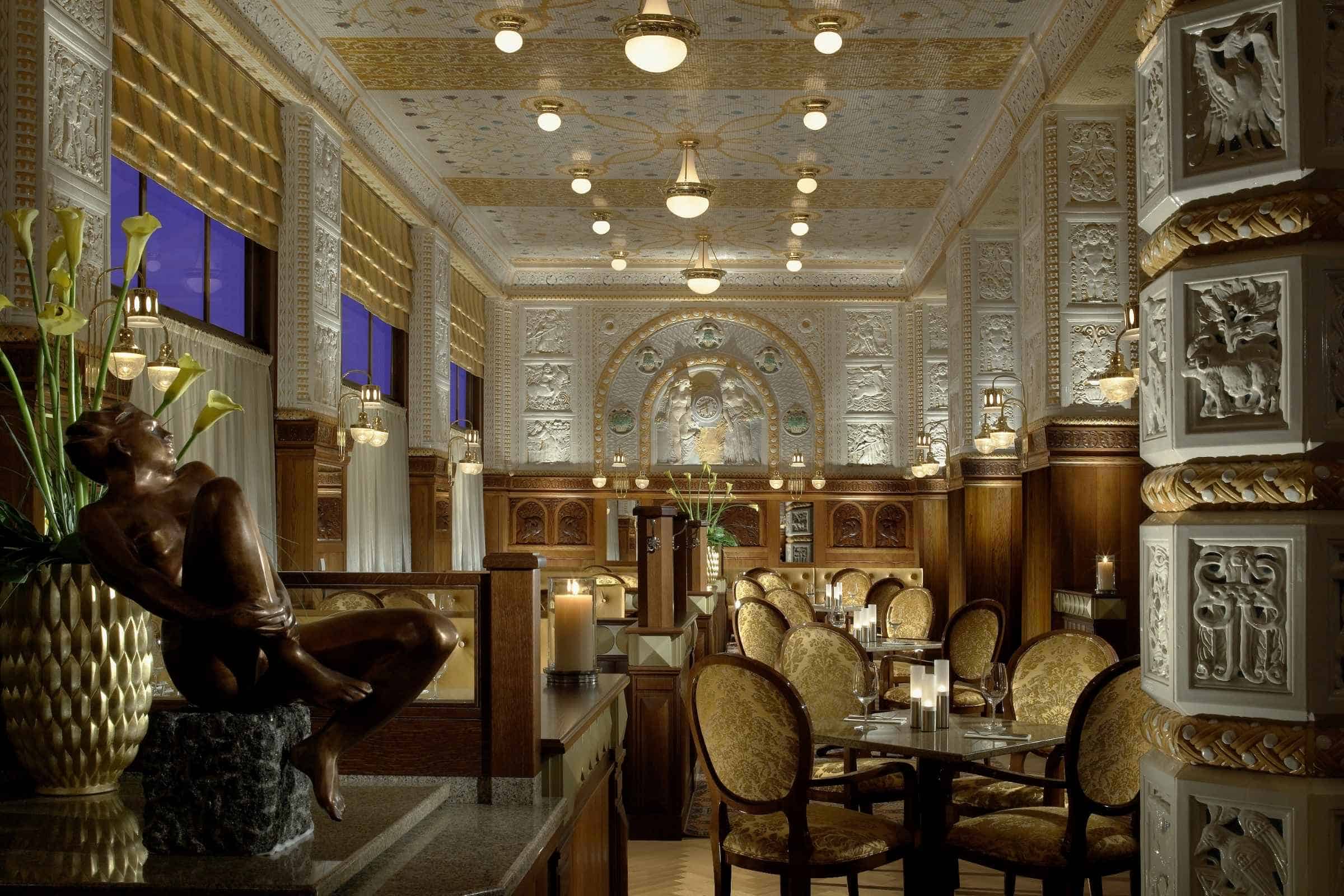 Interior of Café Imperial Prague with mosaic tile ceiling, ceramic tile walls, carved wood wainscoting, Art Deco lighting, sculpture, and ornate frieze