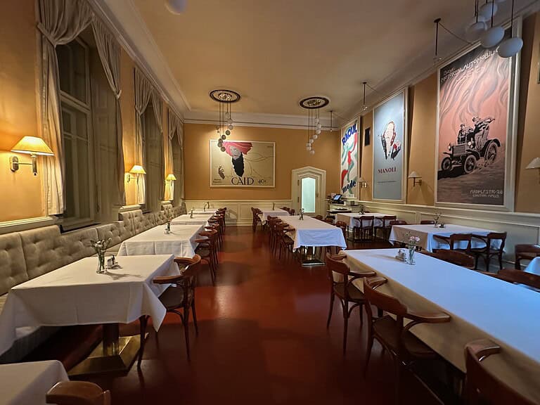 Dining salon at Café Louvre in Prague, with long tables, bentwood chairs, Art Nouveau posters, and soft pendant lighting