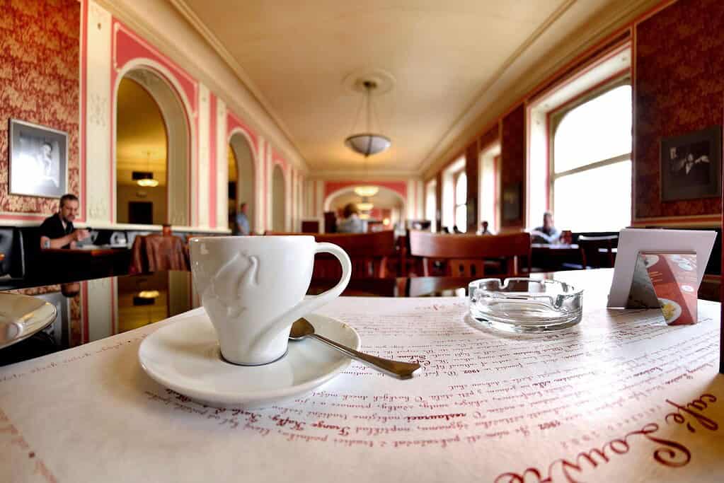 Interior of Café Louvre in Prague with a white coffee cup on a marble-topped table, arched walls, red-and-cream décor, and patrons seated in the background