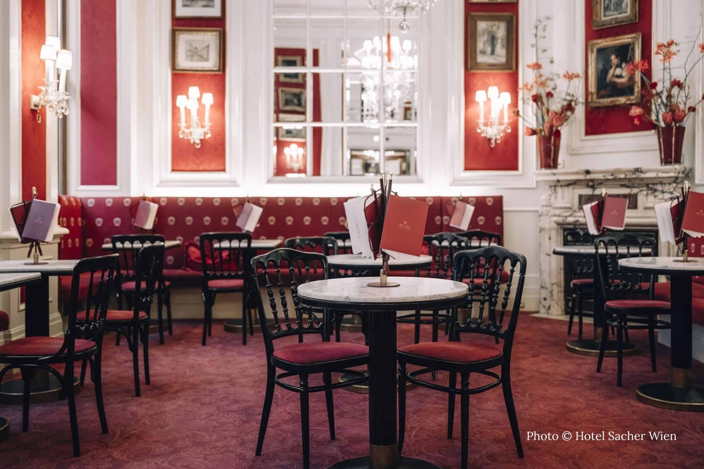 Café Sacher Vienna interior with crystal sconces and mirrored walls Interior of Café Sacher in Vienna with red banquettes, marble tables, and mirrored walls lit by crystal sconces
