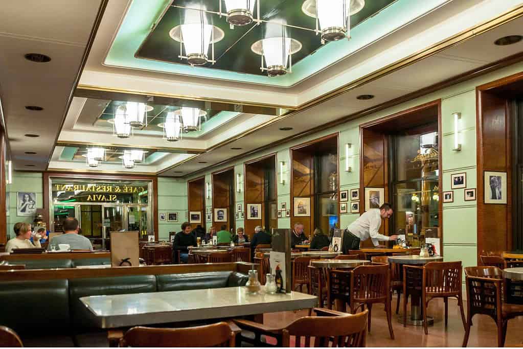 Interior of Café Slavia in Prague with marble-topped tables, wooden chairs, green leather banquettes, and lantern-style ceiling lights, as patrons dine and read
