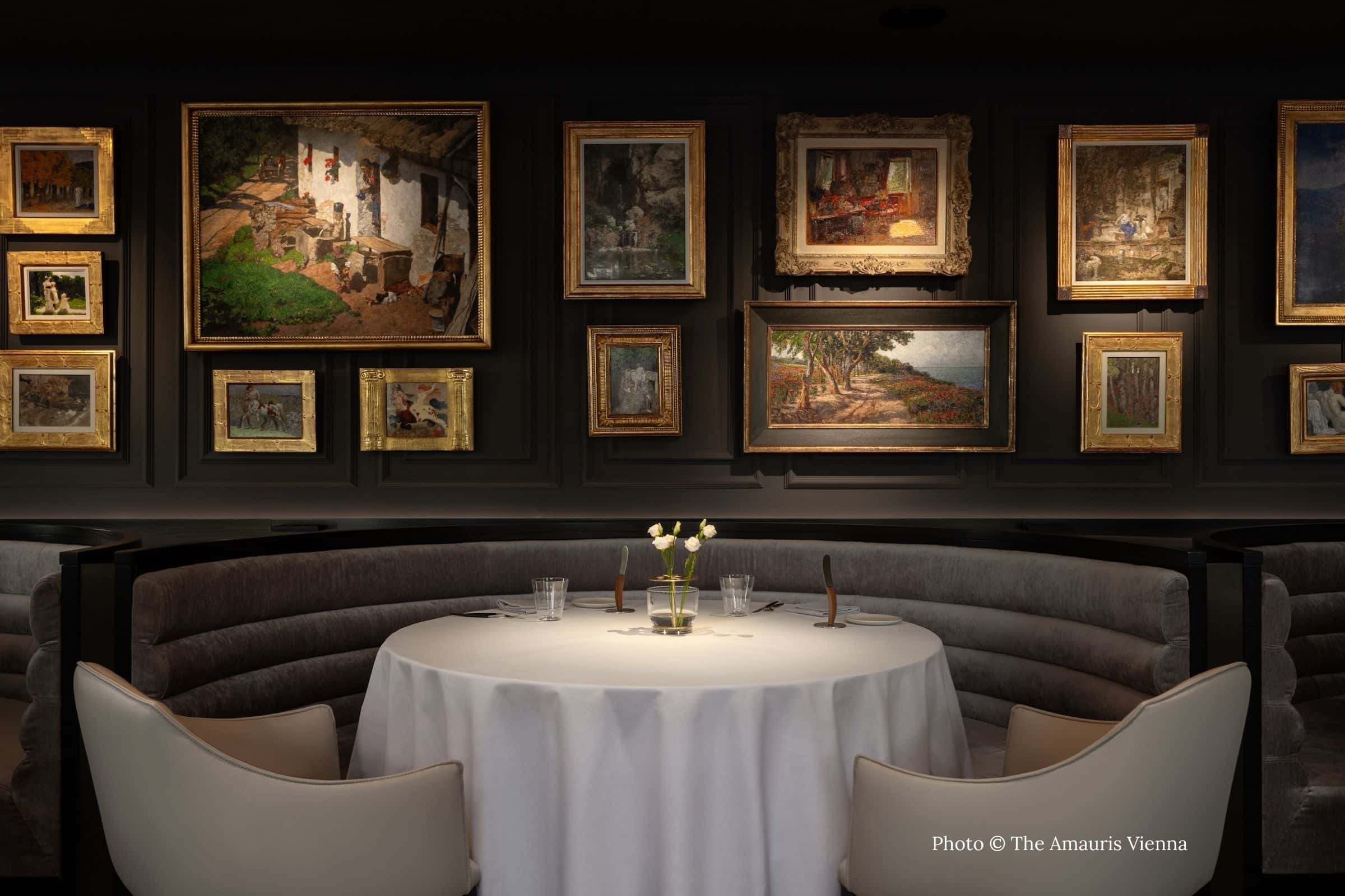 Intimate dining room at Glasswing restaurant in Vienna’s Amauris Hotel, featuring a curved banquette, white-linen table, and a gallery wall of framed paintings