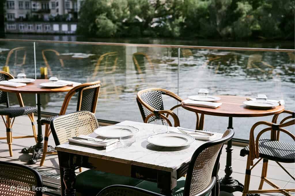 Outdoor and interior views of Mina Berlin along the Spree, from riverside seating and plant-filled dining rooms to Roman-style pizzas finished with mortadella, pistachios, and truffle
