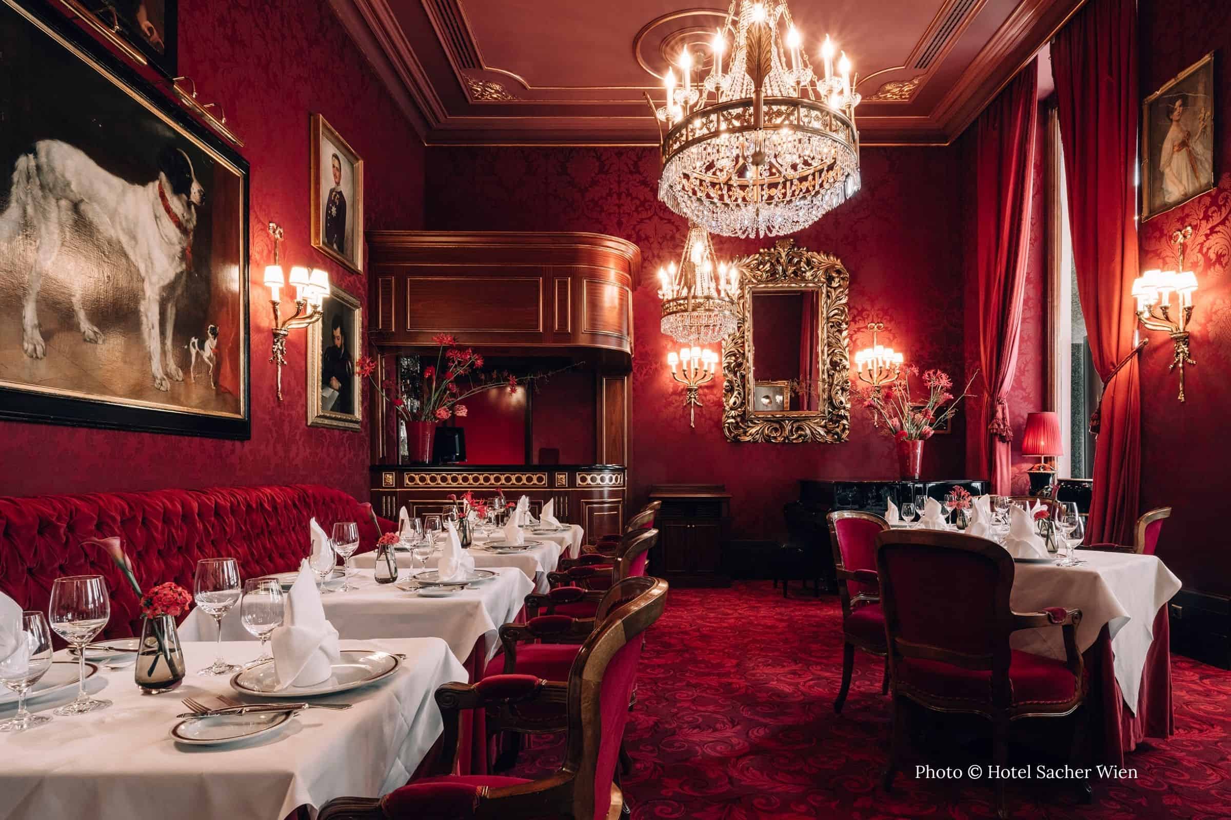 Red velvet dining room at the Rote Bar restaurant in Vienna’s Hotel Sacher, featuring chandeliers, gilded mirrors, damask walls, and white-linen tables