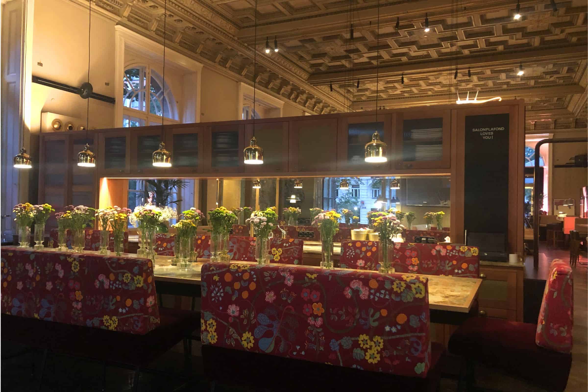 Dining room at Salonplafond restaurant in Vienna’s MAK Museum, featuring floral-upholstered seating, long communal tables, pendant lighting, and historic coffered ceilings