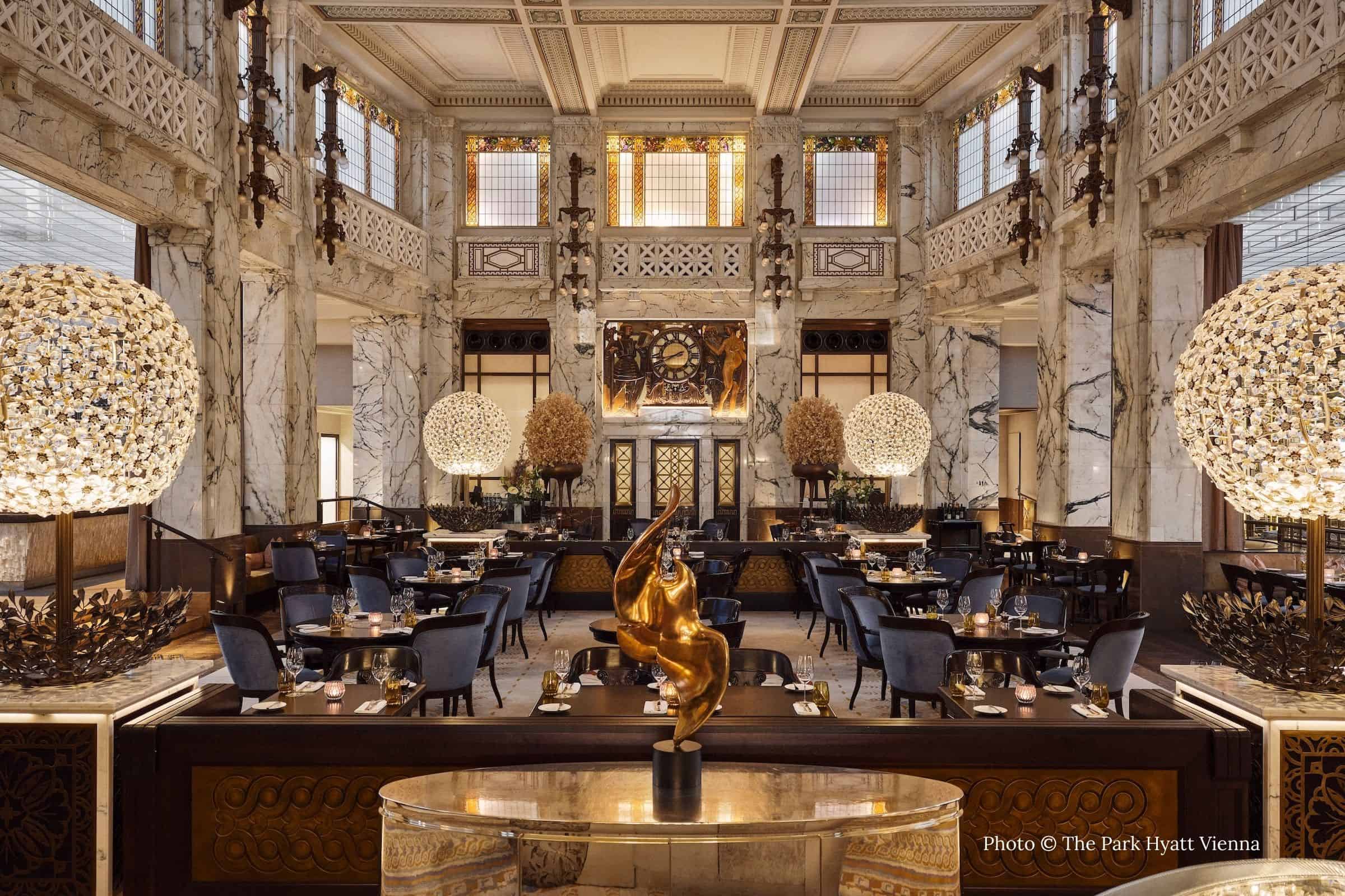 Grand dining room at The Bank restaurant in Vienna’s Park Hyatt Hotel, featuring marble columns, gilded Art Deco details, sculptural lighting, and a central bronze sculpture