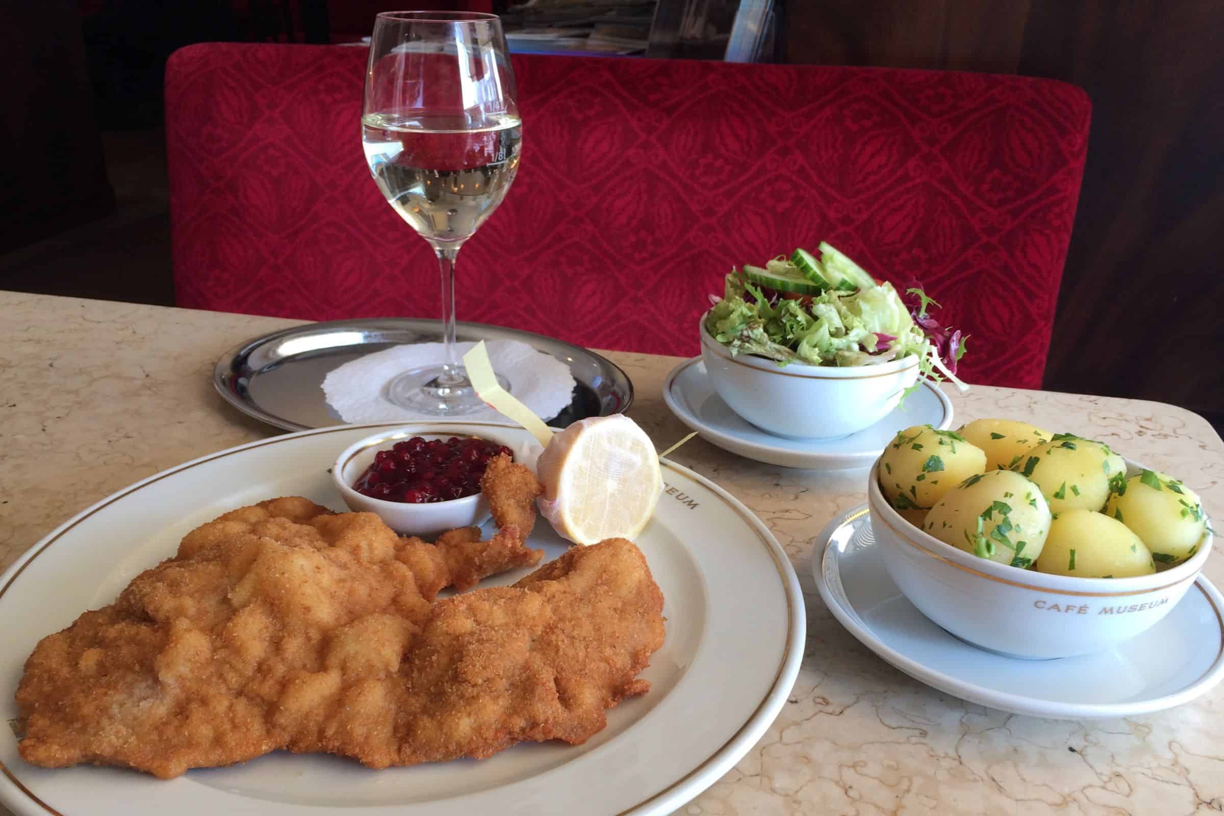 Wiener schnitzel served at Café Museum in Vienna, accompanied by parsley potatoes, green salad, lingonberry sauce, lemon, and a glass of white wine