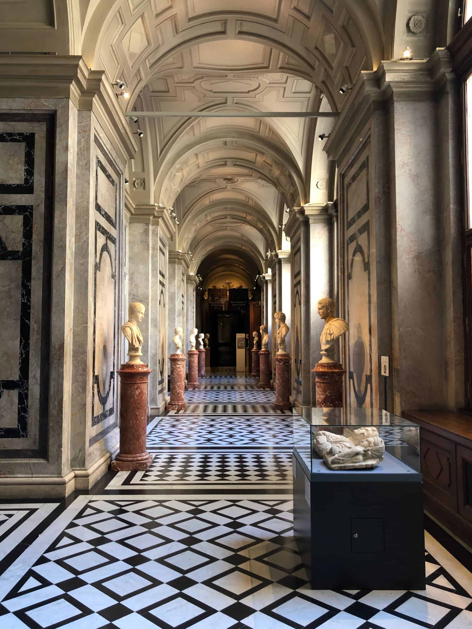 Vaulted corridor in the Kunsthistorisches Museum in Vienna, featuring repeating arches, marble columns, classical sculptures, and a geometric black-and-white floor pattern.