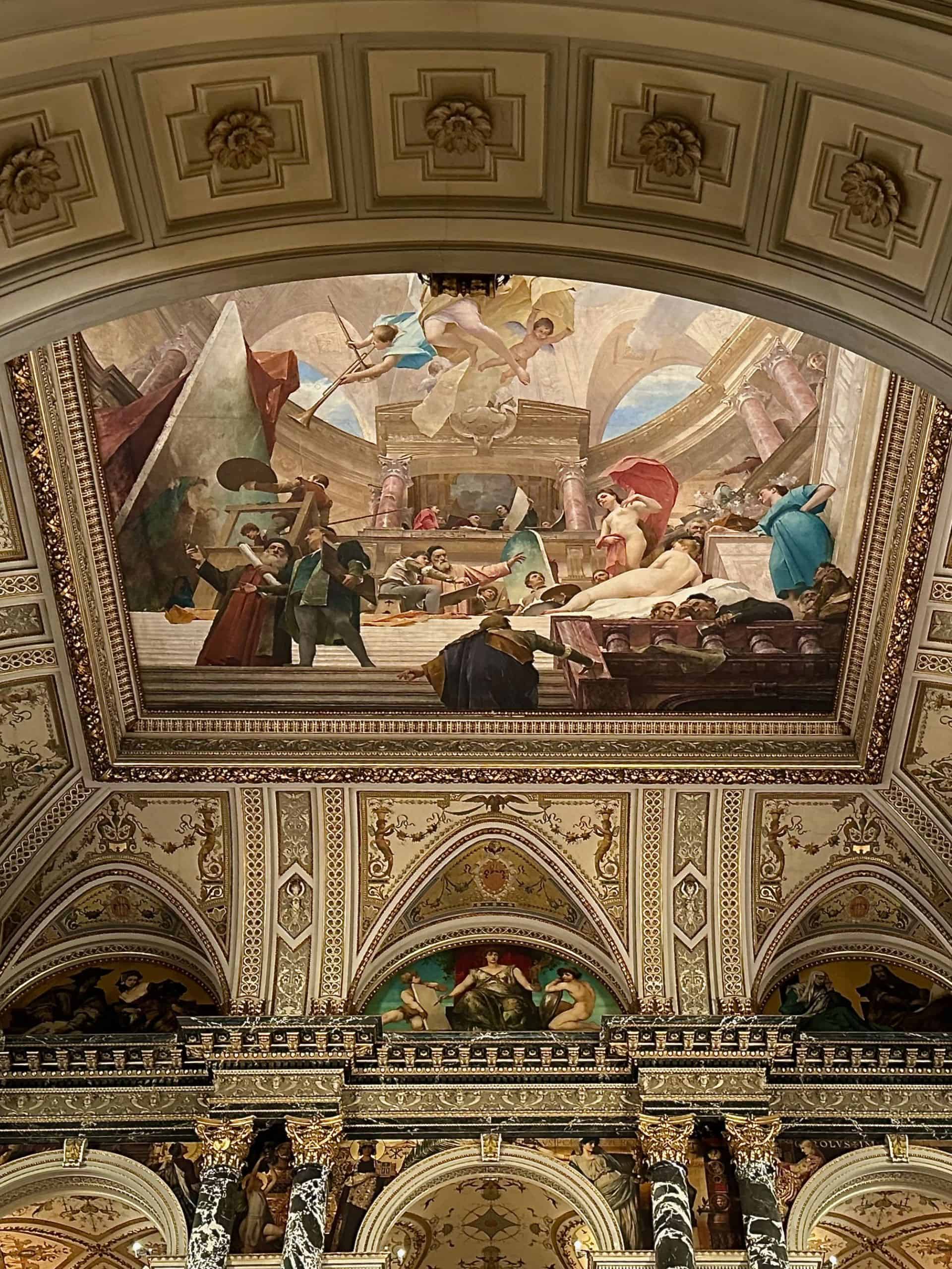 Ceiling frescoes above the grand staircase of the Kunsthistorisches Museum in Vienna, framed by ornate arches, marble columns, and gilded historicist decoration