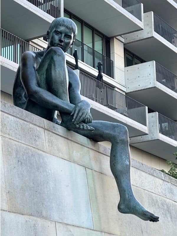 Bronze sculpture along the Spree River in Berlin with modern residential buildings in the background Bronze sculpture of a contemplative boy seated on a stone ledge above the Spree River in Berlin