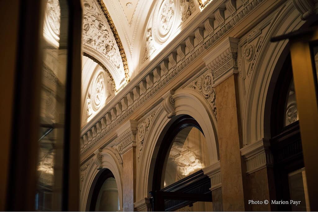 Interior views of Almanac Palais Vienna, a luxury boutique hotel in two restored 19th-century neo-Baroque palaces on Vienna's Ringstrasse, showcasing ornate historic ceilings, modern atrium lobby with abstract art panels and contemporary sculpture, and elegant deluxe room with high gilded ceilings and plush king bed