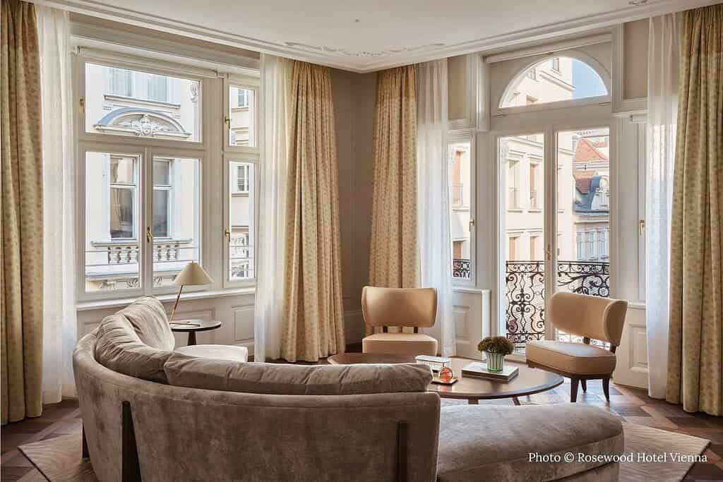 Interior and exterior views of Rosewood Vienna, including rooftop bar seating overlooking the historic dome of the Peterskirche, the neoclassical façade of the former Erste Österreichische Spar-Casse, and a light-filled corner suite with parquet floors
