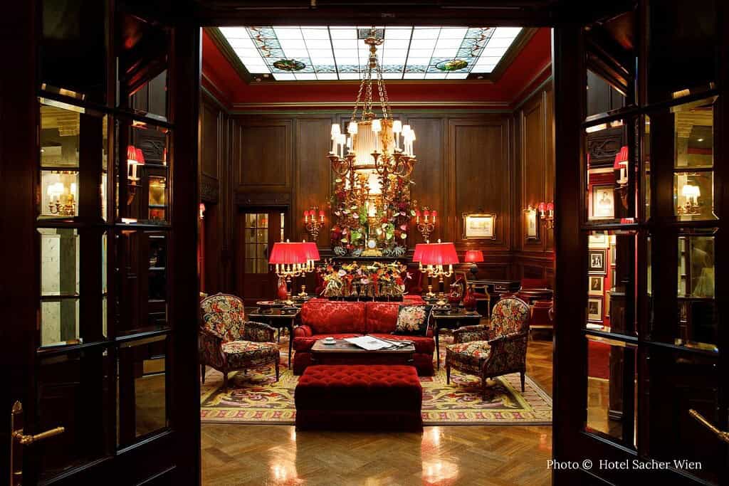 Exterior and interior views of Hotel Sacher Wien, including its illuminated façade opposite the Vienna State Opera, a grand signature suite with red accents, and the wood-paneled lobby salon
