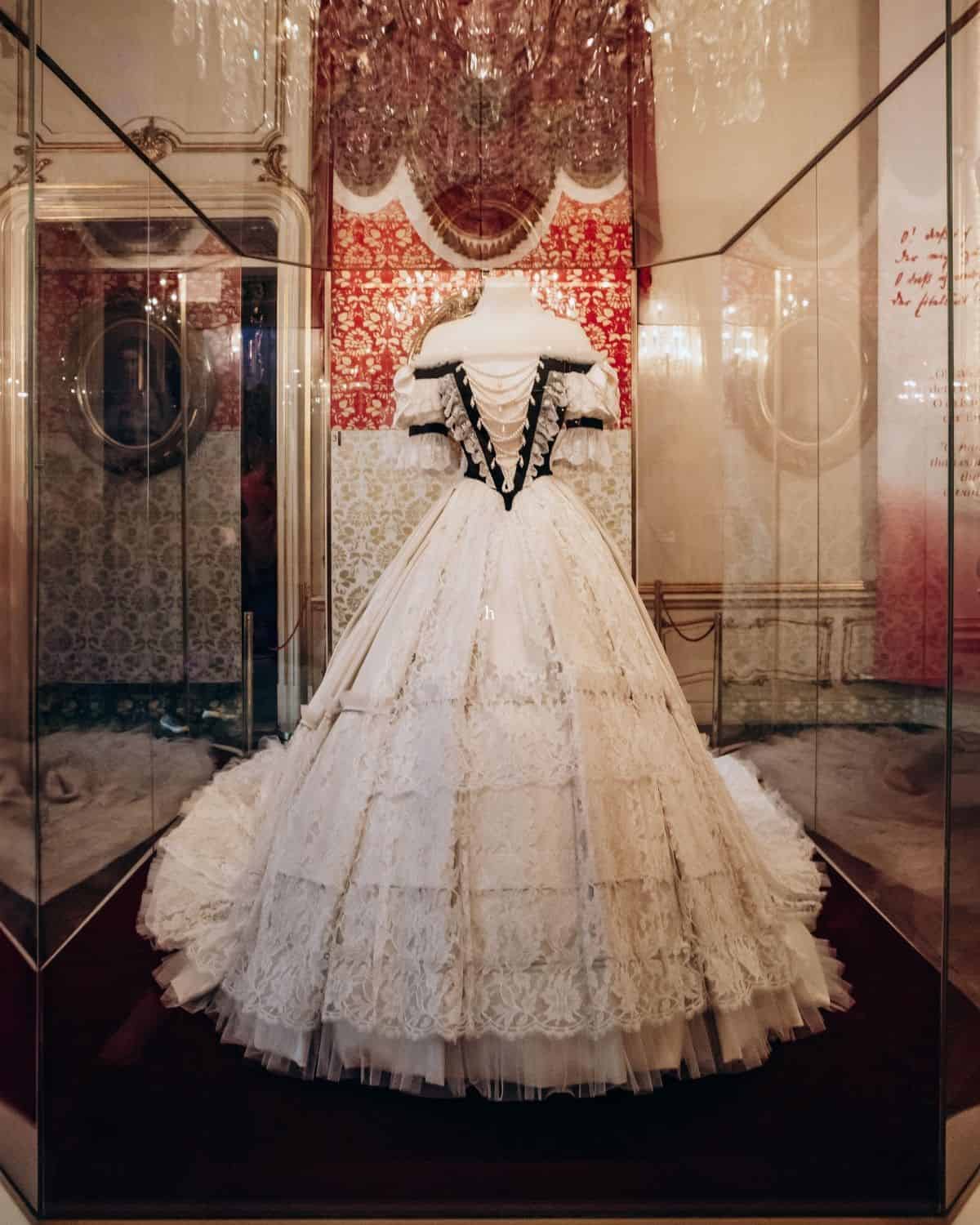 White lace gown associated with Empress Elisabeth displayed in a glass case at the Hofburg in Vienna, with a wide layered skirt and fitted black-trimmed bodice