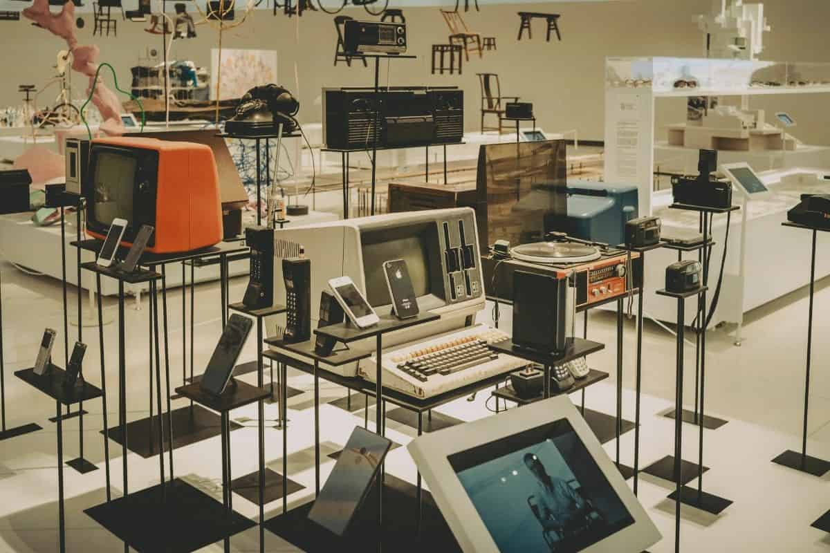 Museum display of vintage televisions, telephones, early computers, radios, cameras, and mobile devices arranged on black stands