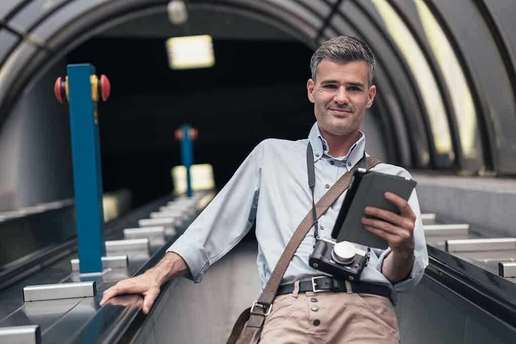 Traveler using a tablet on an escalator with a camera around his neck