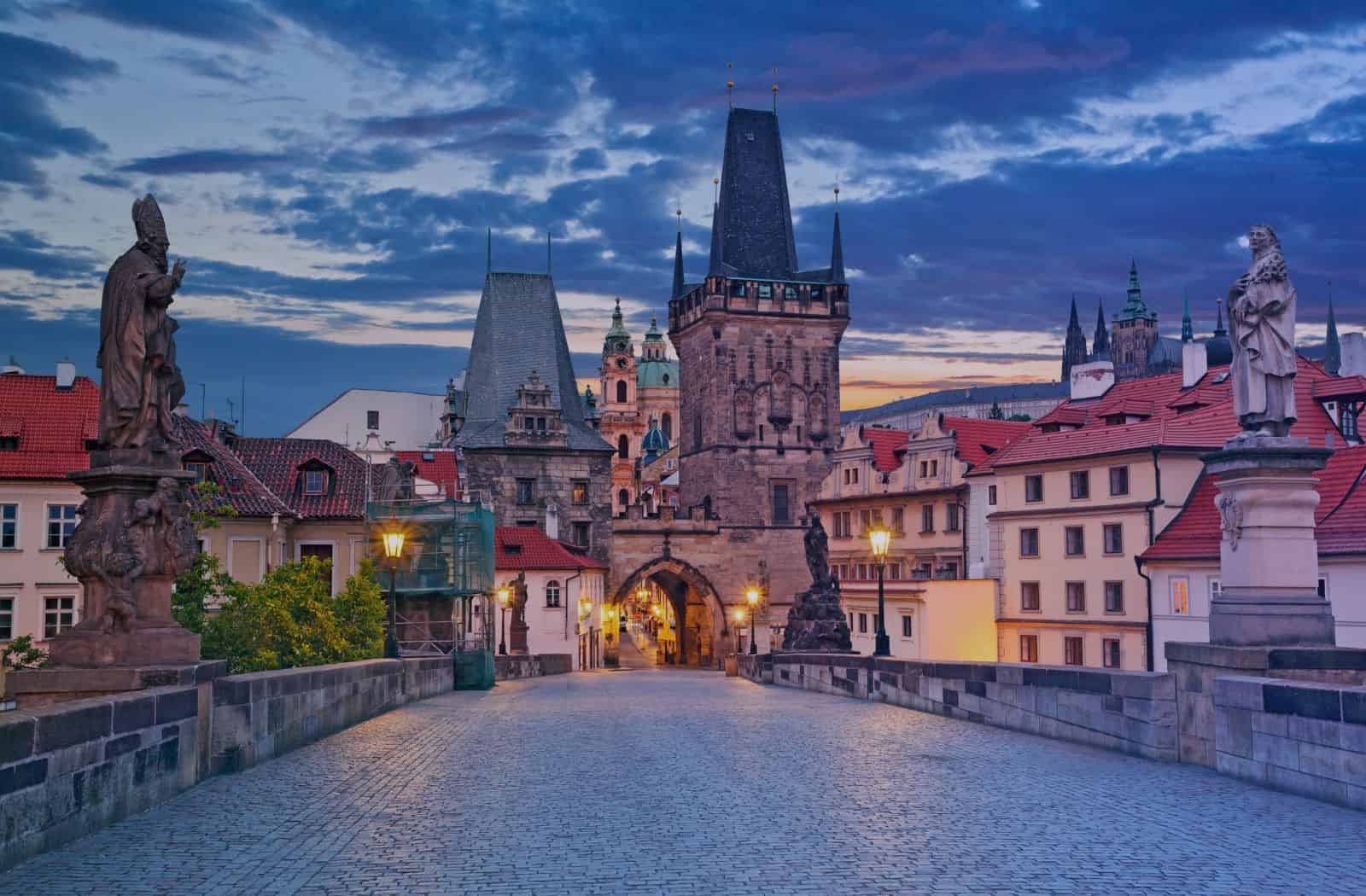 Charles Bridge in Prague at blue hour, facing the Gothic Lesser Town Bridge Tower, with the bridge’s statues, cobblestones, and the spires of St. Vitus Cathedral visible in the distance