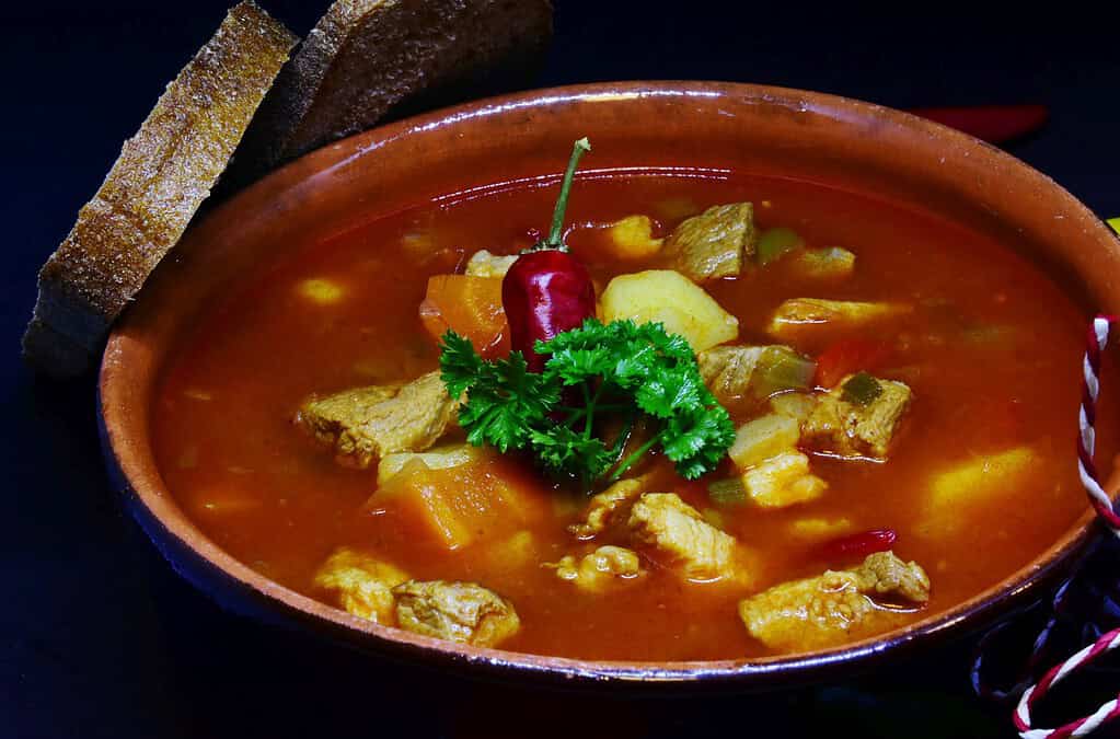 Bowl of Hungarian goulash soup with beef, potatoes, vegetables, parsley, chili pepper, and slices of dark bread