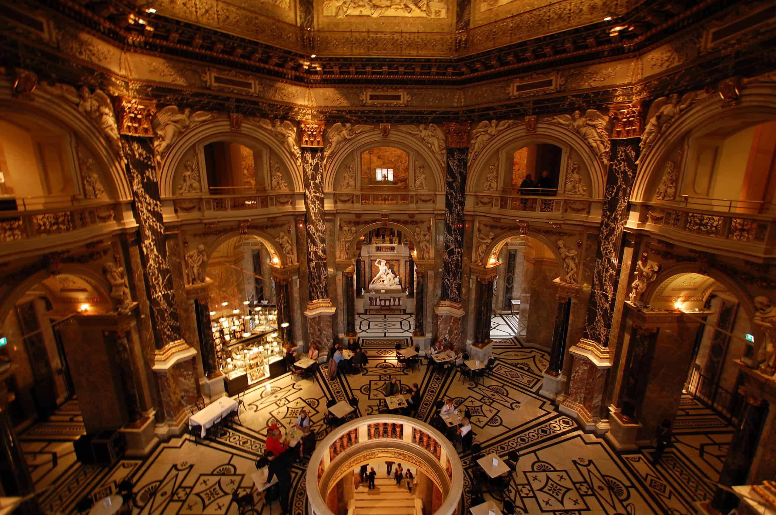 Grand rotunda hall inside the Kunsthistorisches Museum in Vienna, with marble columns, arched galleries, café tables, and visitors below