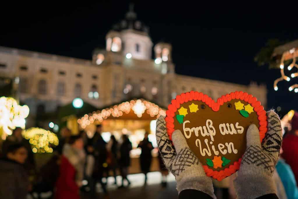 Gloved hands holding a heart-shaped piece of gingerbread at a Christmas Market at Maria-Theresien-Platz with the Natural History Museum in the background