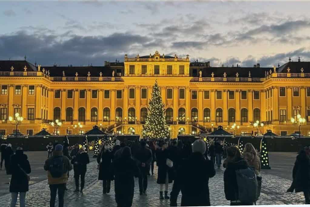 People walking toward Schonbrunn Palace in Vienna at dusk as white Christmas lights sparkle